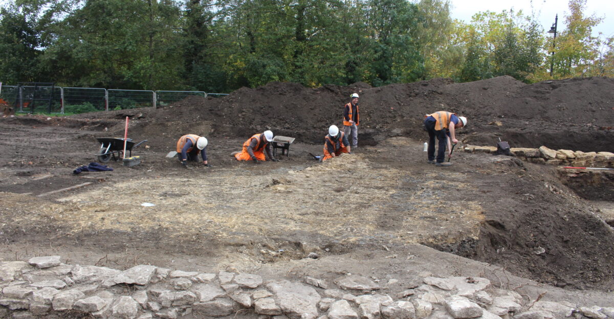 Five archaeologists cleaning a footpath surface with trowels