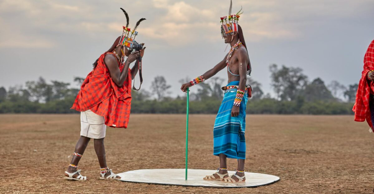 Two Maasai men in traditional attire one taking a photo with a DSLR camera outdoors