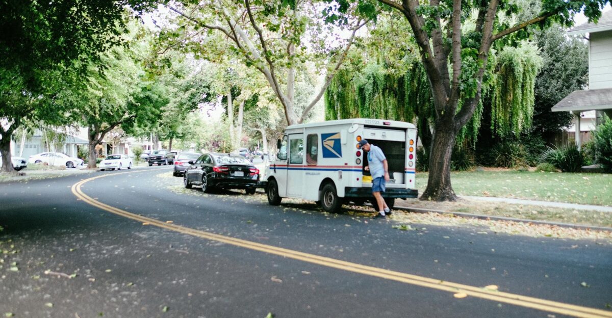 A US mail carrier retrieves packages from a postal van on a peaceful tree-lined street