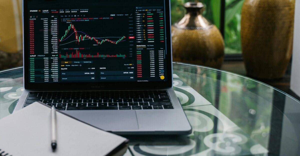 Laptop displaying cryptocurrency stocks and graphs on a glass table with a notepad