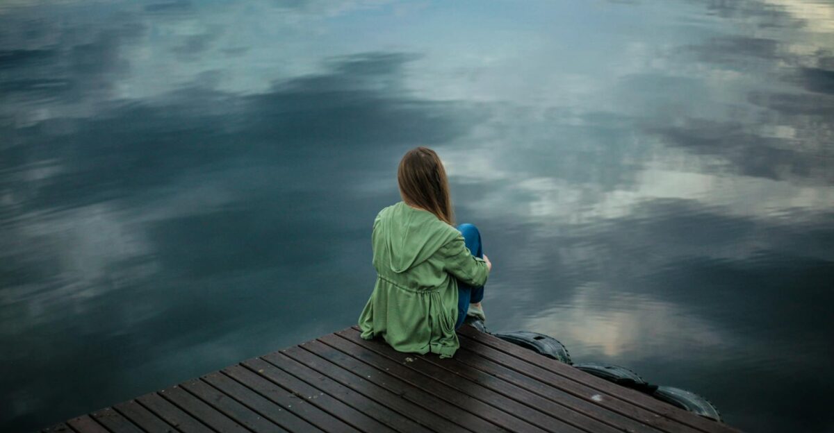 A woman sits on a wooden dock reflecting by a calm lake under a cloudy sky