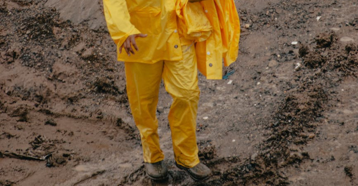 A construction worker in yellow rain gear and helmet holding a clipboard stands on a muddy site