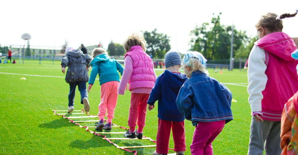 Children enjoy an outdoor activity on a grassy field stepping over a ladder