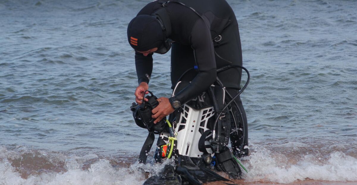 A diver in a wetsuit prepares photography equipment on a beach shoreline