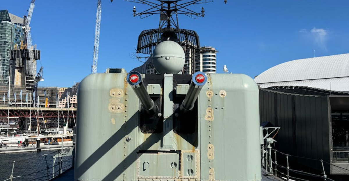 A naval turret mounted on a warship docked in a sunny harbor surrounded by cityscape
