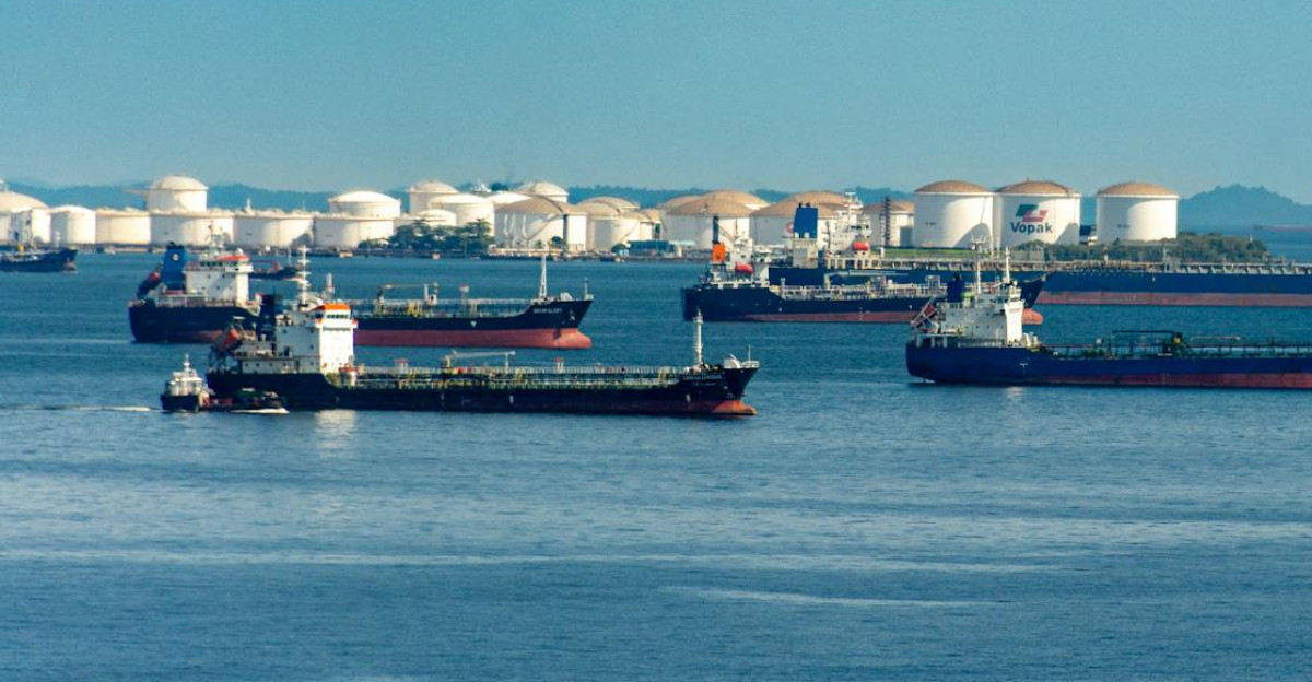 A fleet of cargo ships docked near oil storage tanks along a serene coastline with a clear blue sky above