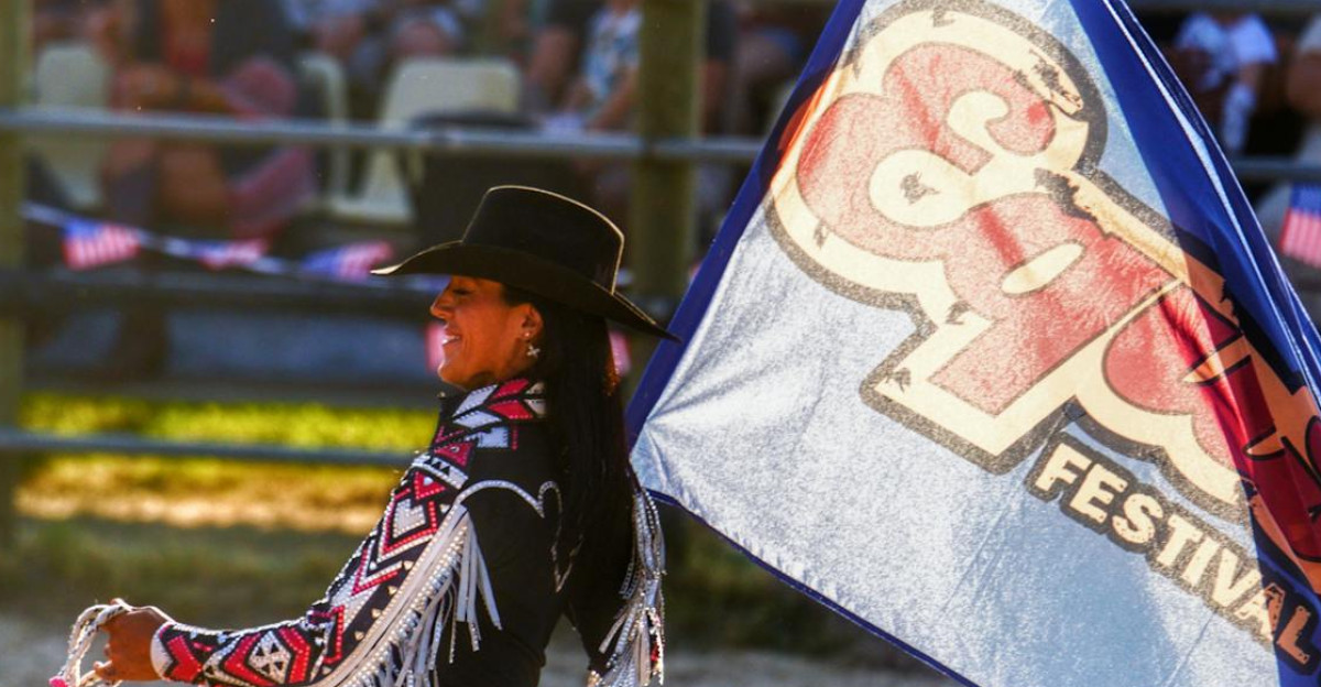 A cowgirl carrying a festival flag rides a horse at a sunny rodeo event