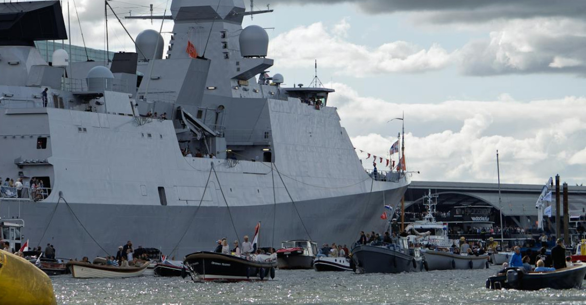 A large naval warship anchored near a modern cityscape under dramatic cloudy skies
