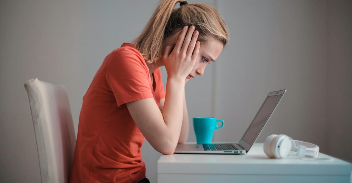 A woman sits indoors at a table looking concerned while using a laptop with headphones and a coffee mug nearby