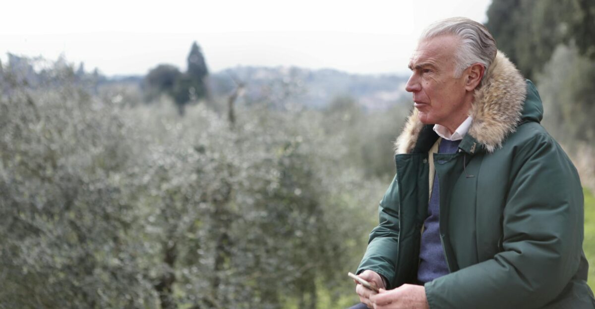 Elderly man in green fur-lined jacket holding phone while sitting outdoors