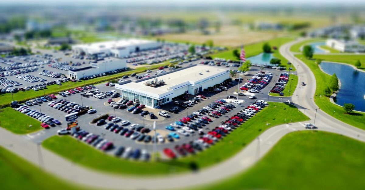 Aerial high-angle view of a bustling car dealership surrounded by parked cars in a green landscape