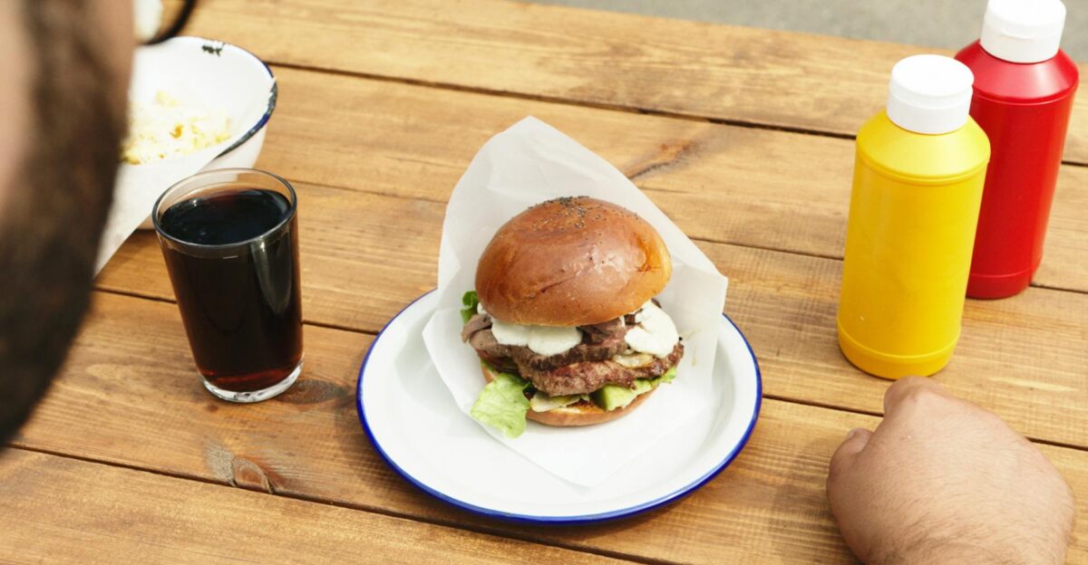 Close-up of a cheeseburger with lettuce accompanied by soda and condiments on a wooden table