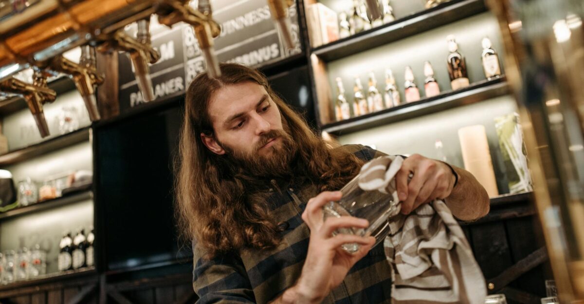 A bearded bartender cleaning a glass with a towel in a cozy bar setting showcasing bartending skills