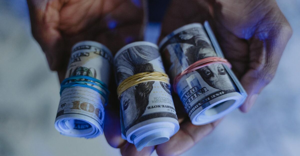 Close-up of hands holding rolled US dollar banknotes secured with rubber bands set against a blue-lit background