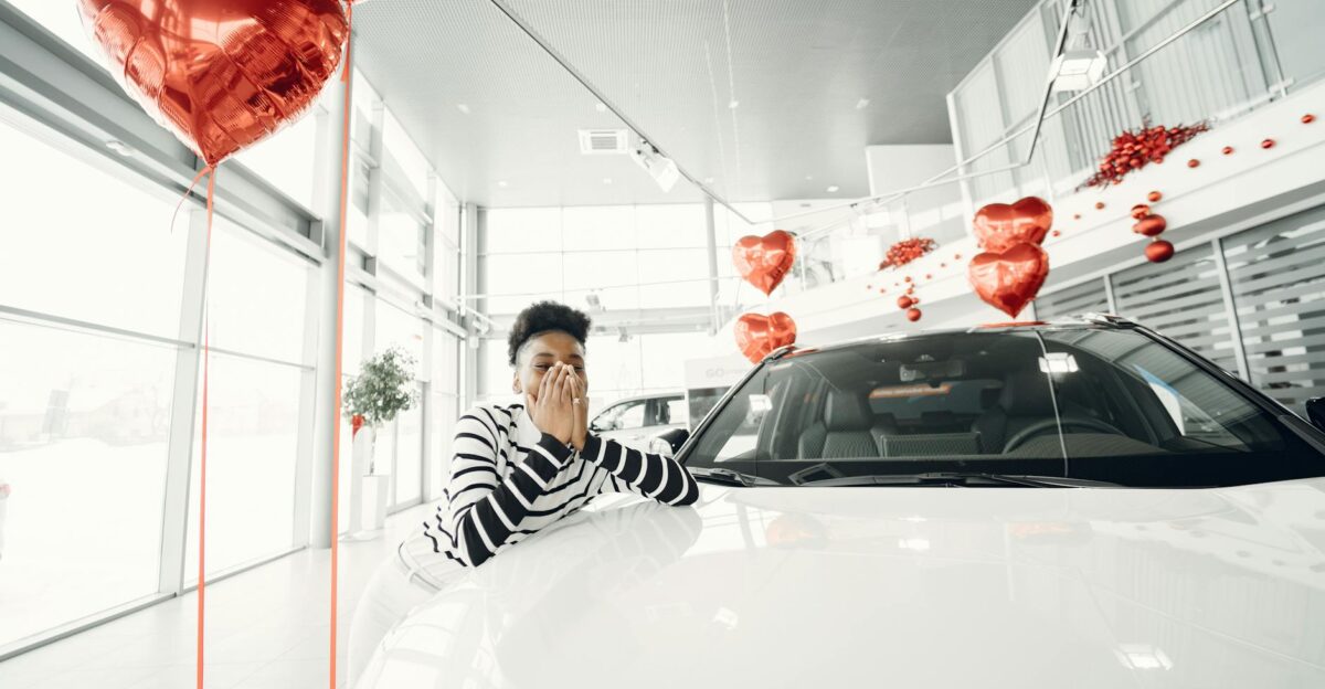 A young woman expresses excitement at a car dealership surrounded by heart-shaped red balloons