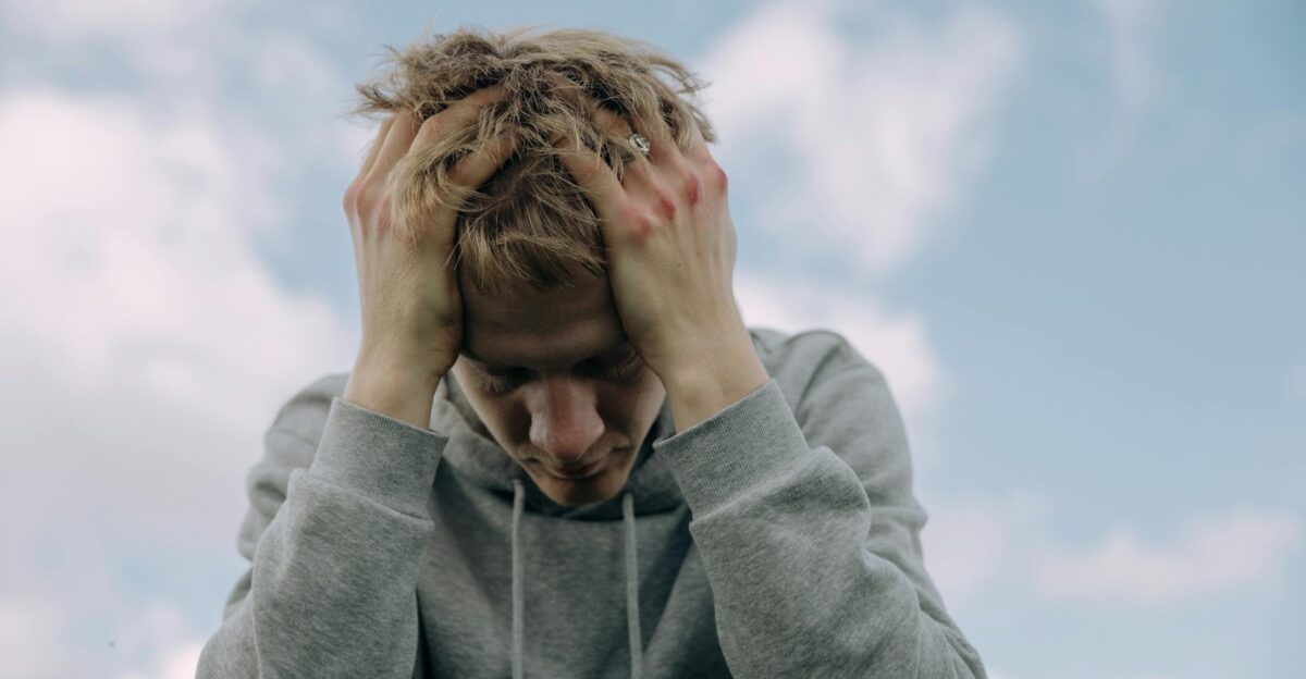 Young man in gray hoodie holds head in frustration set against cloudy sky