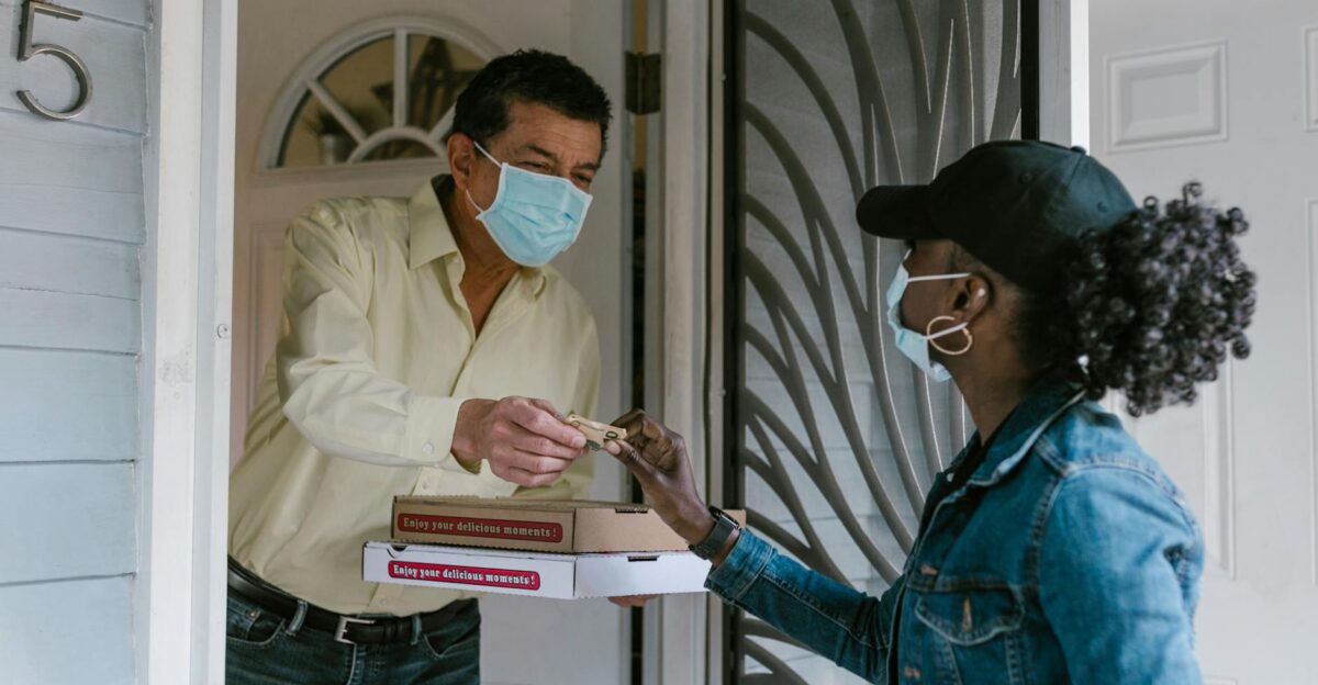 A delivery person hands a pizza to a masked customer at the door showcasing safe delivery services