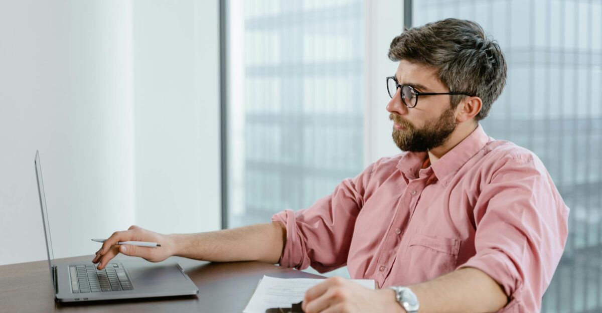 A focused businessman in a pink shirt uses a laptop at his office desk with cityscape views