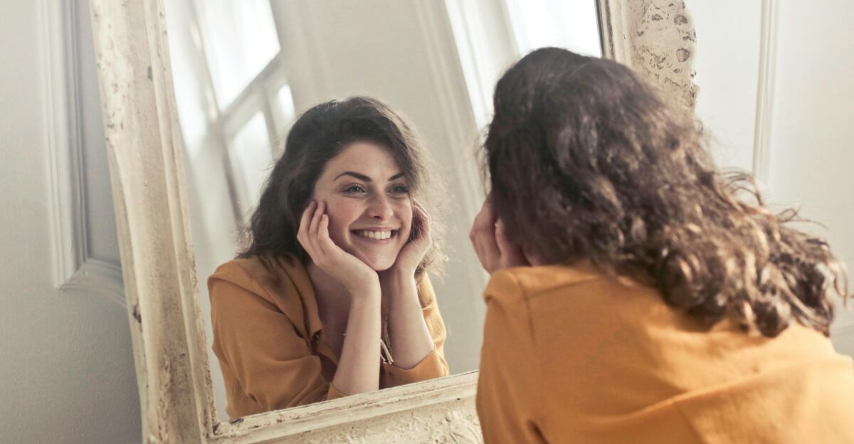 A cheerful woman smiles at her reflection in a vintage-style mirror exuding positivity and warmth
