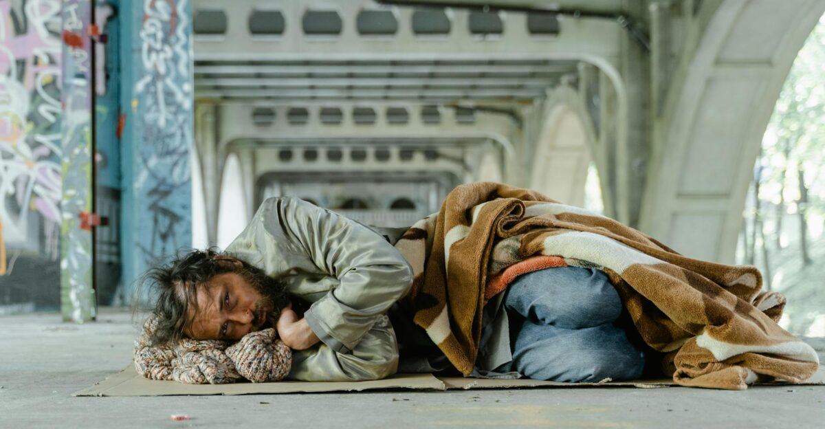 A homeless man lies under an urban bridge wrapped in a blanket symbolizing poverty and survival