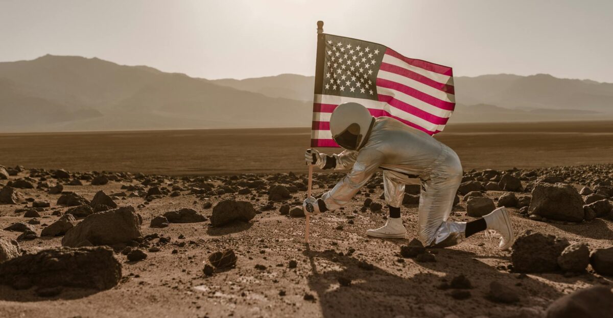 Astronaut in spacesuit planting American flag on rocky desert terrain under the sunlight