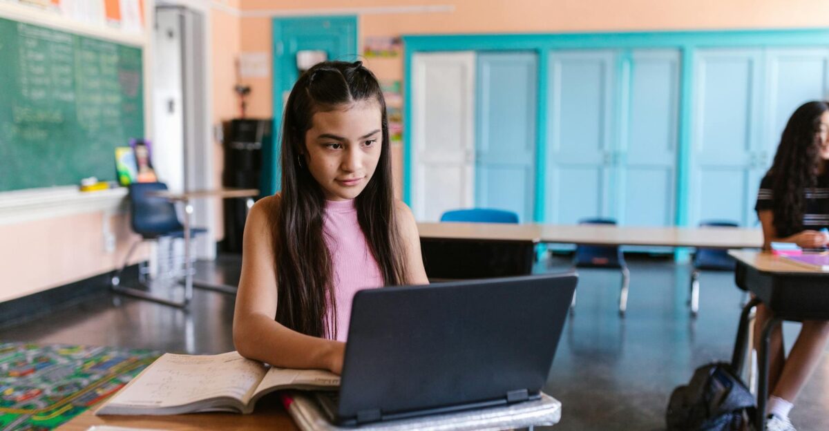 A focused young girl using a laptop for schoolwork in a bright classroom environment