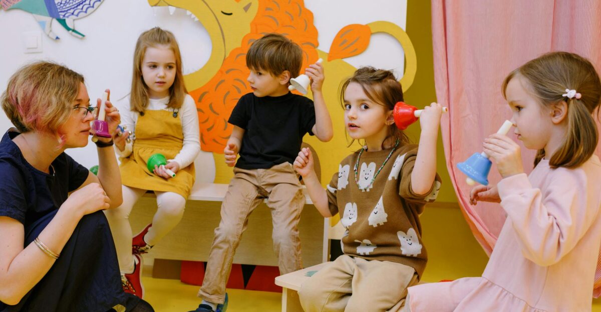 Young children playing musical bells in a colorful classroom setting with a teacher