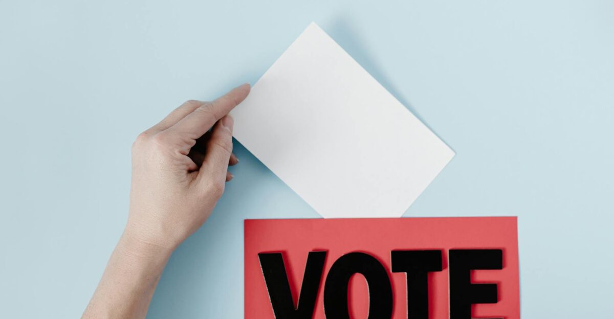 A hand places a ballot into a red box with the word VOTE against a blue background
