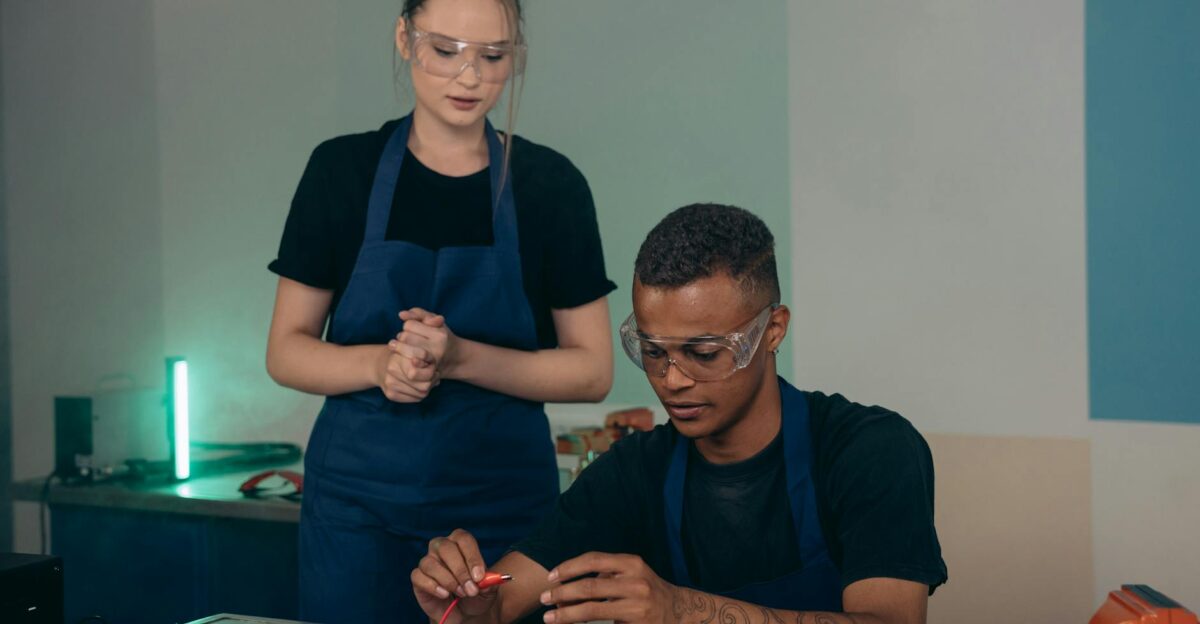 Two colleagues working with electronics in an indoor workshop wearing aprons and safety goggles
