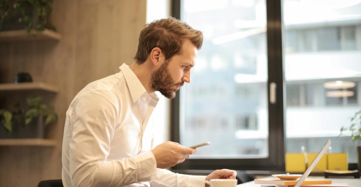 Focused businessman working on laptop while checking smartphone in modern office