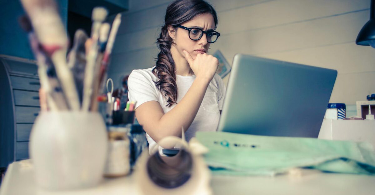 Young woman with glasses deeply focused on a laptop surrounded by art supplies in a home office