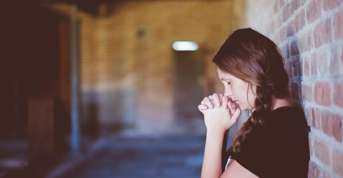 woman praying while leaning against brick wall