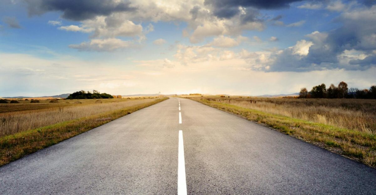 A long straight road stretches through fields under a dramatic cloudy sky