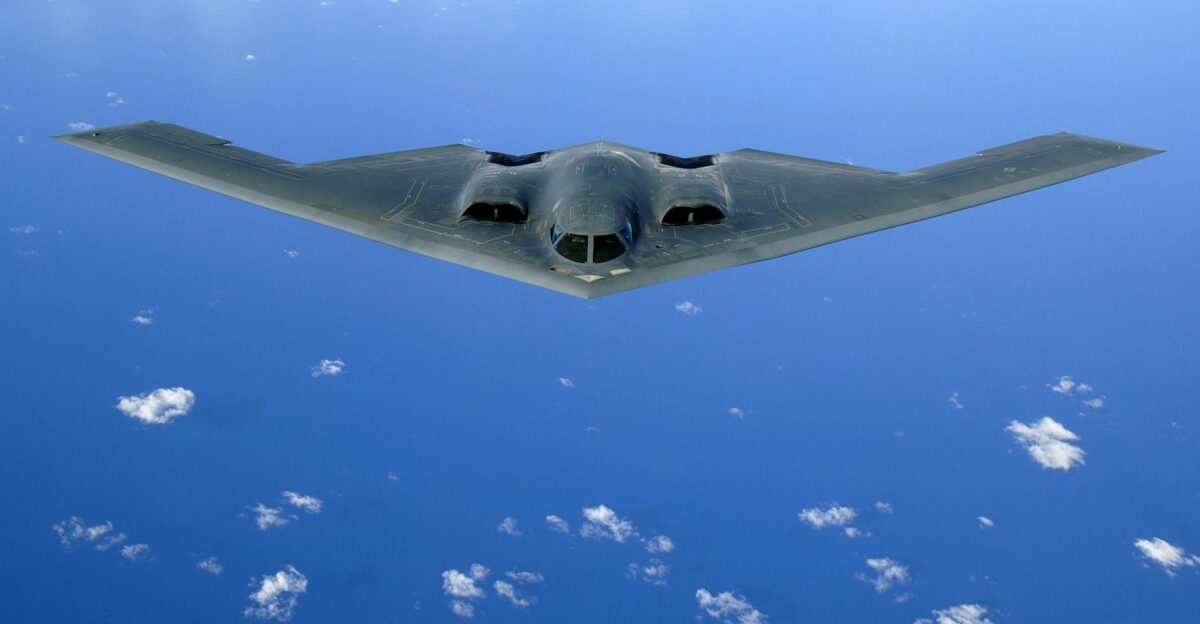 A stealth bomber aircraft flying over a clear blue ocean with scattered clouds