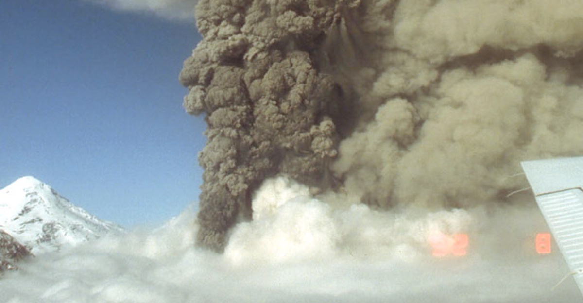 Aerial view, looking north, of the eruption column from the Crater Peak vent, Mount Spurr volcano. A light-tan cloud ascending from pyroclastic flows is visible at right. The 3,374-m (11,070 ft)-high summit lava dome complex of Mount Spurr is visible at left.