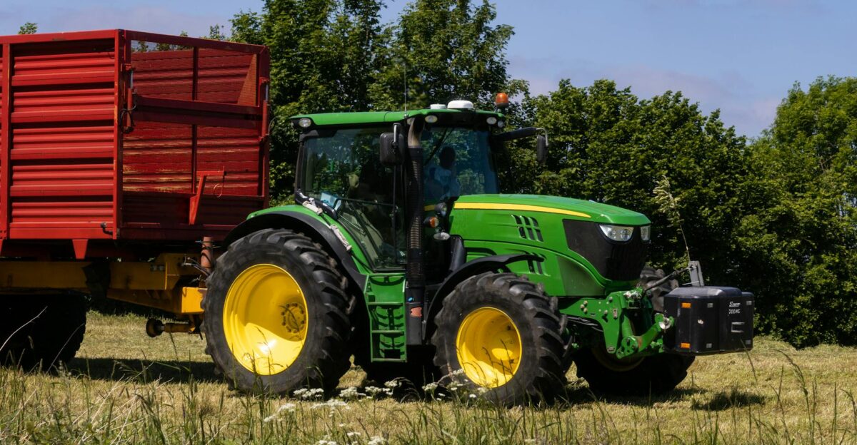 A green tractor with a yellow wheel pulls a red trailer in a sunny countryside field