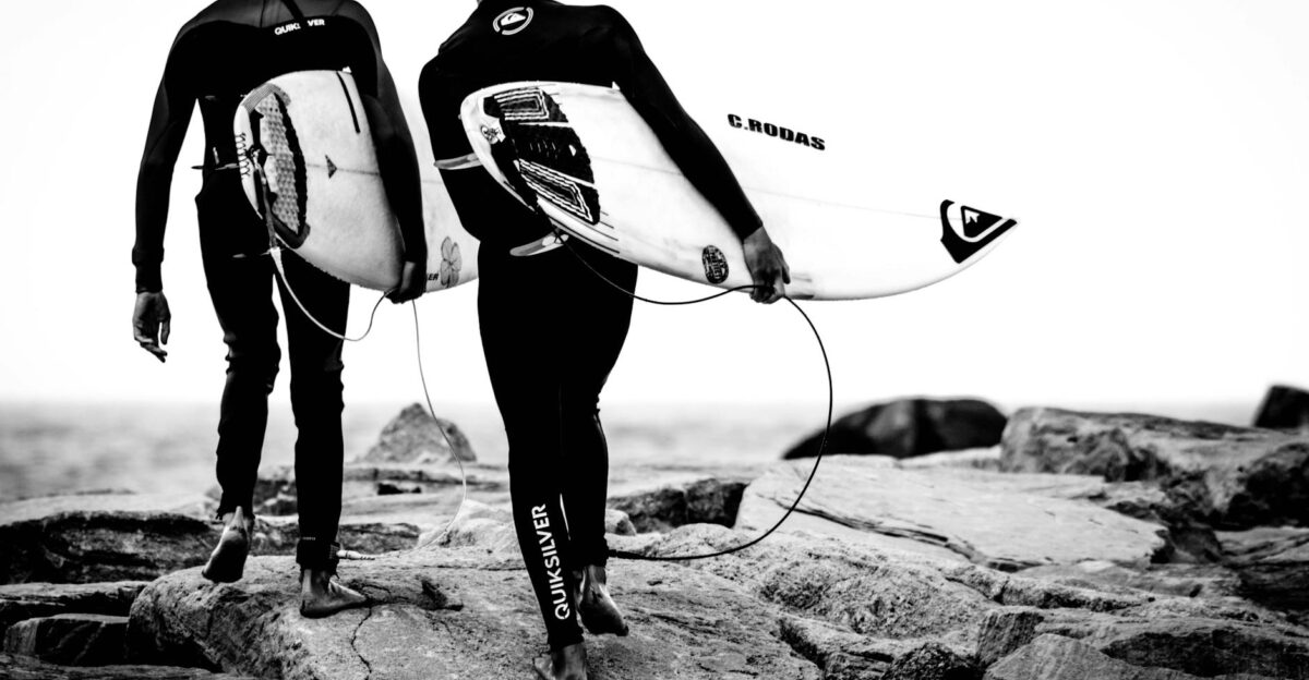 Two surfers walking on rocks with surfboards by the sea in black and white