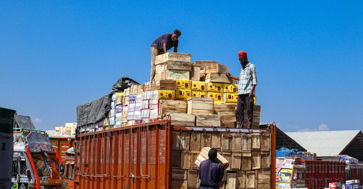 Three men loading wooden boxes on a truck under a clear blue sky in Sopore