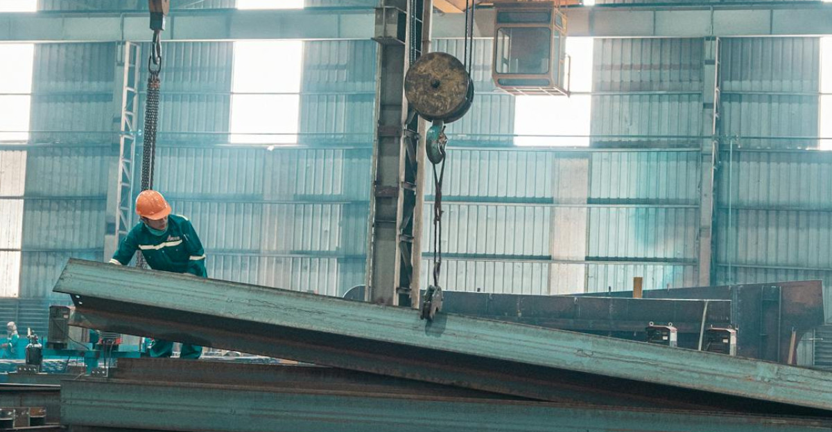 A worker in a helmet manages steel beams in a large factory setting
