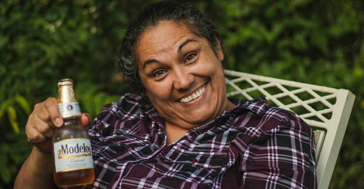 Happy woman smiling broadly while holding a Modelo beer in a garden setting