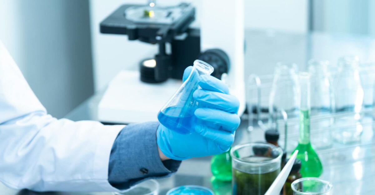 Scientist in gloves analyzing blue liquid in a laboratory setting with microscope and glassware