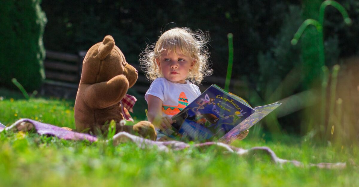 Young child reading a book outdoors with a teddy bear enjoying a sunny day on the grass