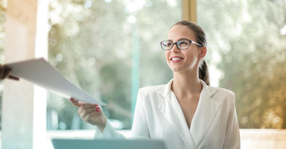 Low angle of successful female executive manager in classy style sitting at table with laptop in contemporary workplace and passing documents to colleague
