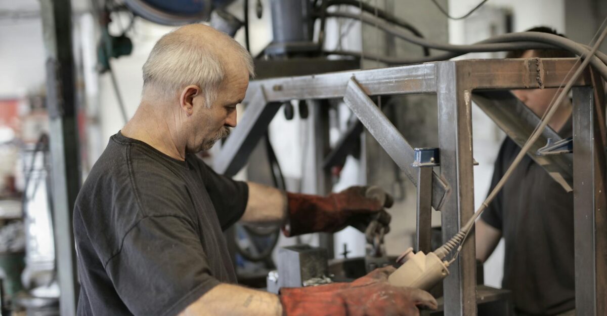 Side view of senior white hair craftsman in heavy duty gloves dealing with machine in workshop