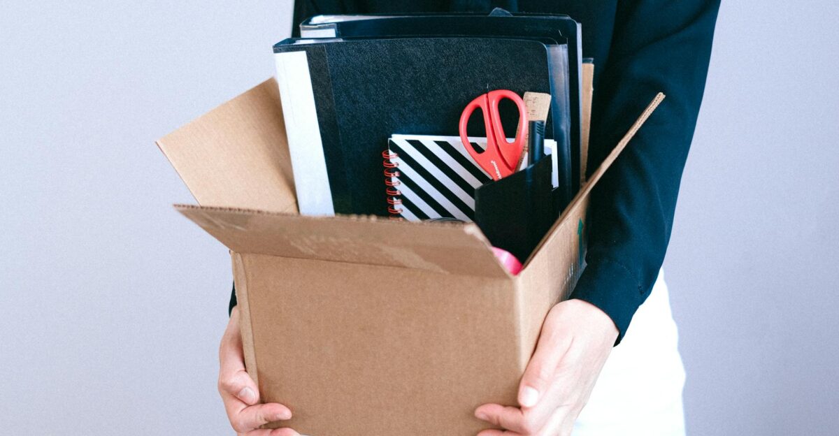 Woman carrying cardboard box with office items like scissors and notebooks