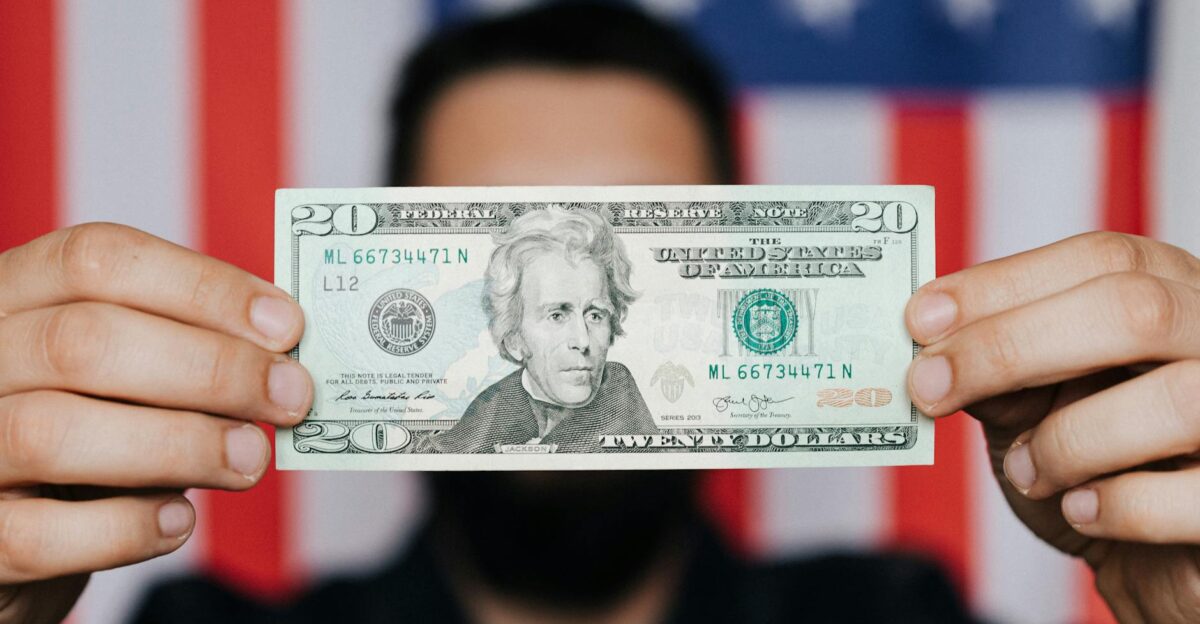 Close-up of a man holding a 20-dollar bill with an American flag blurred in the background symbolizing finance and patriotism