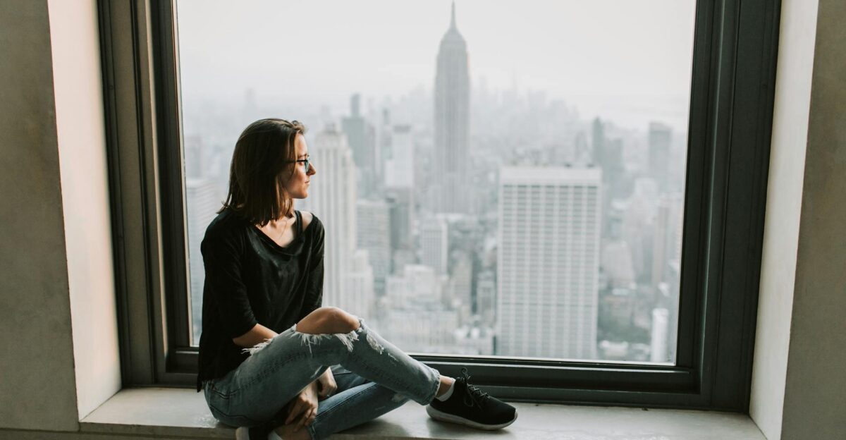 A woman sits on a windowsill admiring the New York City skyline with the Empire State Building in view
