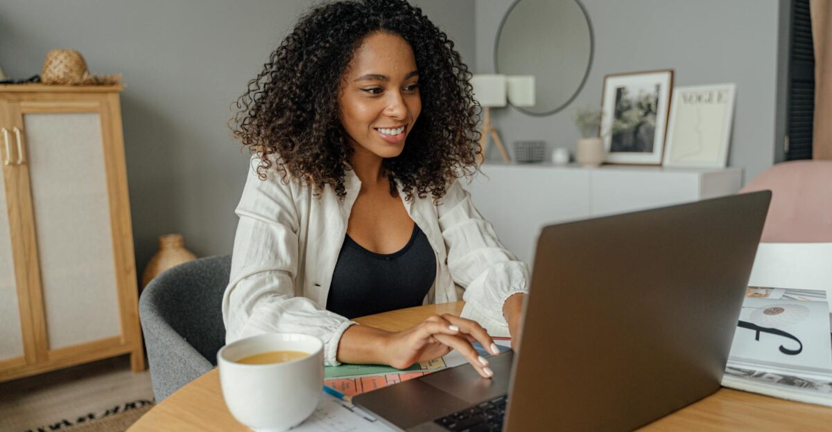 A woman sits at a round table working on a laptop with a coffee cup nearby in a cozy home office setting