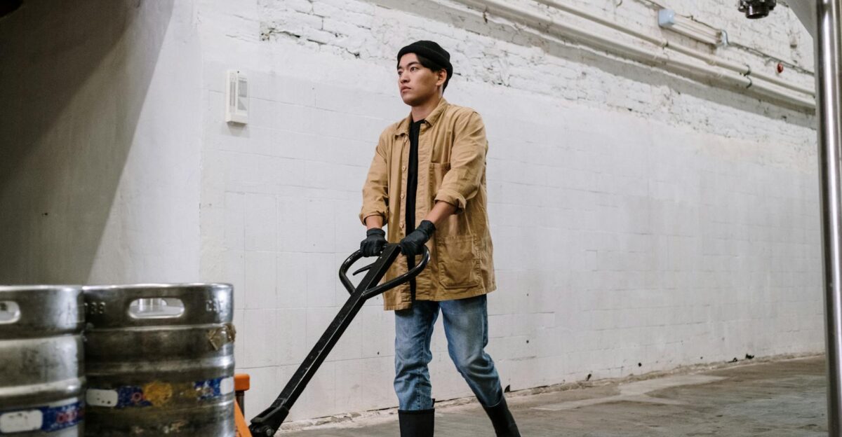 Young man in brewery moving barrels with hand truck Modern industrial setting