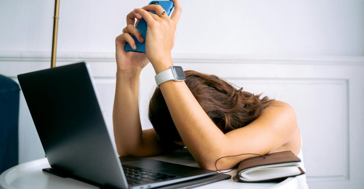 A woman overwhelmed by work resting head on table with laptop phone and smartwatch